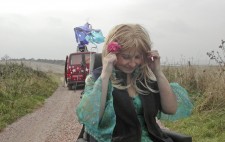 An a country lane with grassy banks, and against a looming sky, Liz Crow is dressed in the same green dress and wig, a flower in her hair and a navy blue gilet added for warmth. In the background is the red van with its spray painted decoration, Liz's wheelchair perched high on its rooftop, with fabric billowing in the wind.