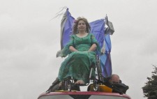 Against a looming sky, Liz Crow, perched on the roof of the van, her wheelchair strapped in place, is dressed in the same green dress and wig, fabric flowing from the back of her chair. Lying flat along the roof of the van are two of the crew holding tightly to the chair for additional anchorage.