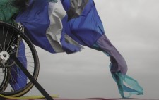 The rear wheel of a wheelchair is strapped to the roof of a red ban, a great train of blue, grey and purple ripstock fabric billowing in the wind against a solid grey sky.